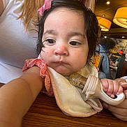 Zylah is registered to the contest to win money with this photo: baby, child, teething_toy, pink_bow, bib, table, wood, restaurant, people, woman, curly_hair, light_fixture, casual_clothing, indoor, dining, hand, skin, face, expression, seated