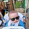 baby, child, sunglasses, swing, porch, wood, hair, blue_clothing, bib, outdoor, greenery, cardboard_boxes, fence, reflection, seat, rope, casual, expression, face, sunny