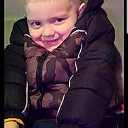 Dom is registered to the contest to win money with this photo: child, boy, jacket, puffy_jacket, smile, face, hands, sitting, indoor, casual_clothing, young, cute, portrait, hairstyle, playful, person, fun, warm_clothing, expression, cozy