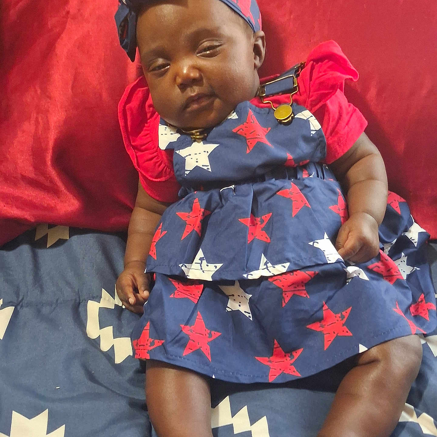 Janiyah is registered to the contest to win money with this photo: baby, infant, child, sleepy, blue_dress, red_shirt, headband, bed, blanket, patterned_clothing, stars, indoors, skin, legs, feet, hand, cute, resting, portrait, person
