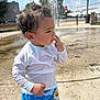 toddler, child, curly_hair, white_shirt, colorful_shorts, water_fountain, outdoor, sunny, concrete, side_profile, finger_on_lips, playful, summer_clothing, park, daytime, thinking, casual_wear, water_splash, urban, young_kid