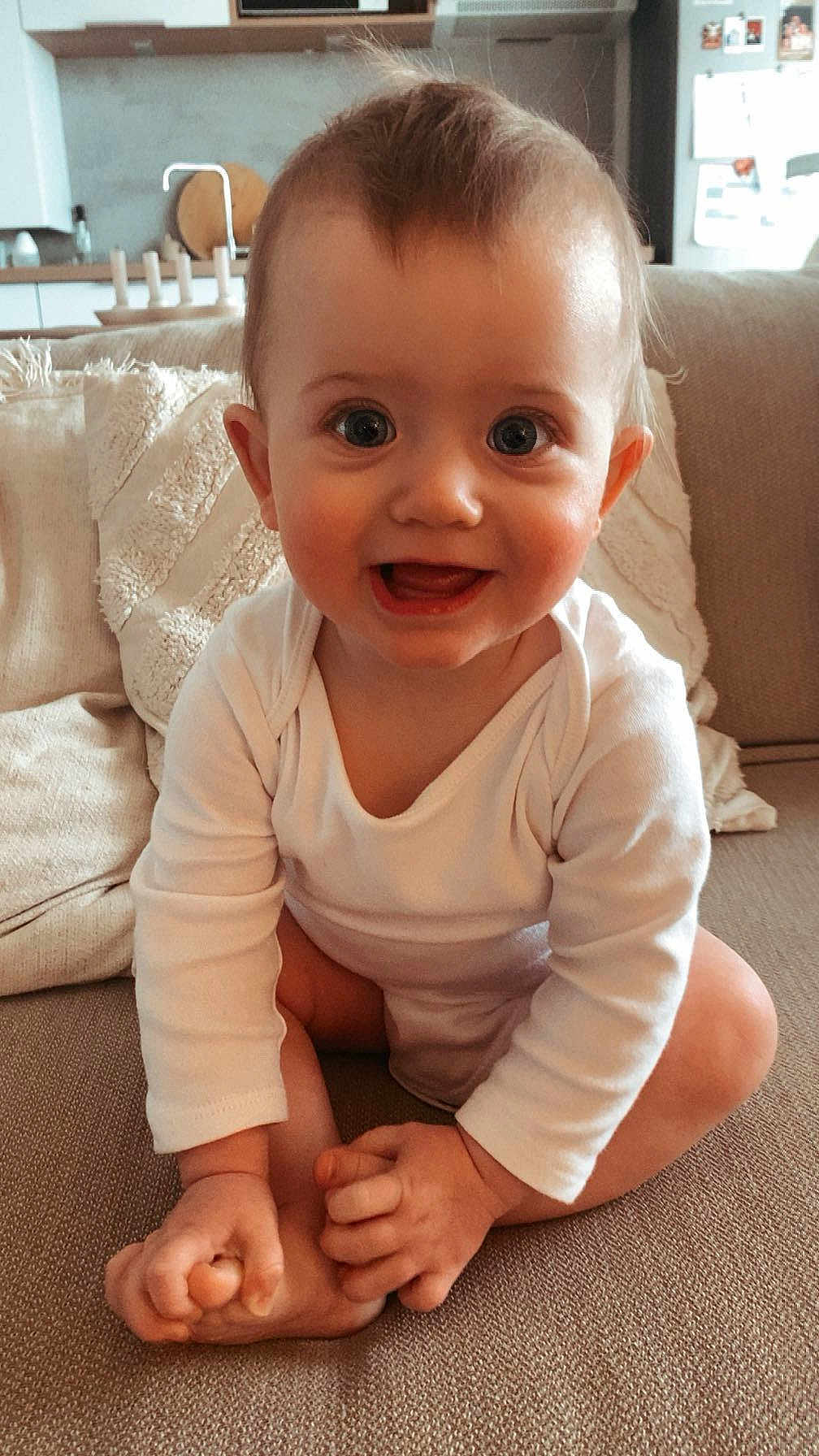 Ama participe au concours pour gagner de l'argent avec cette photo : baby, child, infant, smiling, sitting, hands, feet, couch, home, indoors, white_clothing, cute, happy, portrait, face, person, furniture, living_room, skin, young