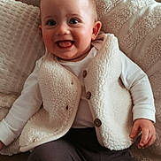 Ama participe au concours pour gagner de l'argent avec cette photo : baby, child, smiling, happy, sitting, couch, vest, white_clothing, dark_pants, face, portrait, indoor, cozy, cute, infant, person, home, soft_texture, buttoned_clothing, tongue_out