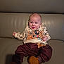 baby, infant, smile, sweater, cartoon_print, fuzzy_booties, pants, leather_sofa, couch, portrait, indoor, happy, cute, hands, eyes, head, sitting, skin, bokeh, cozy