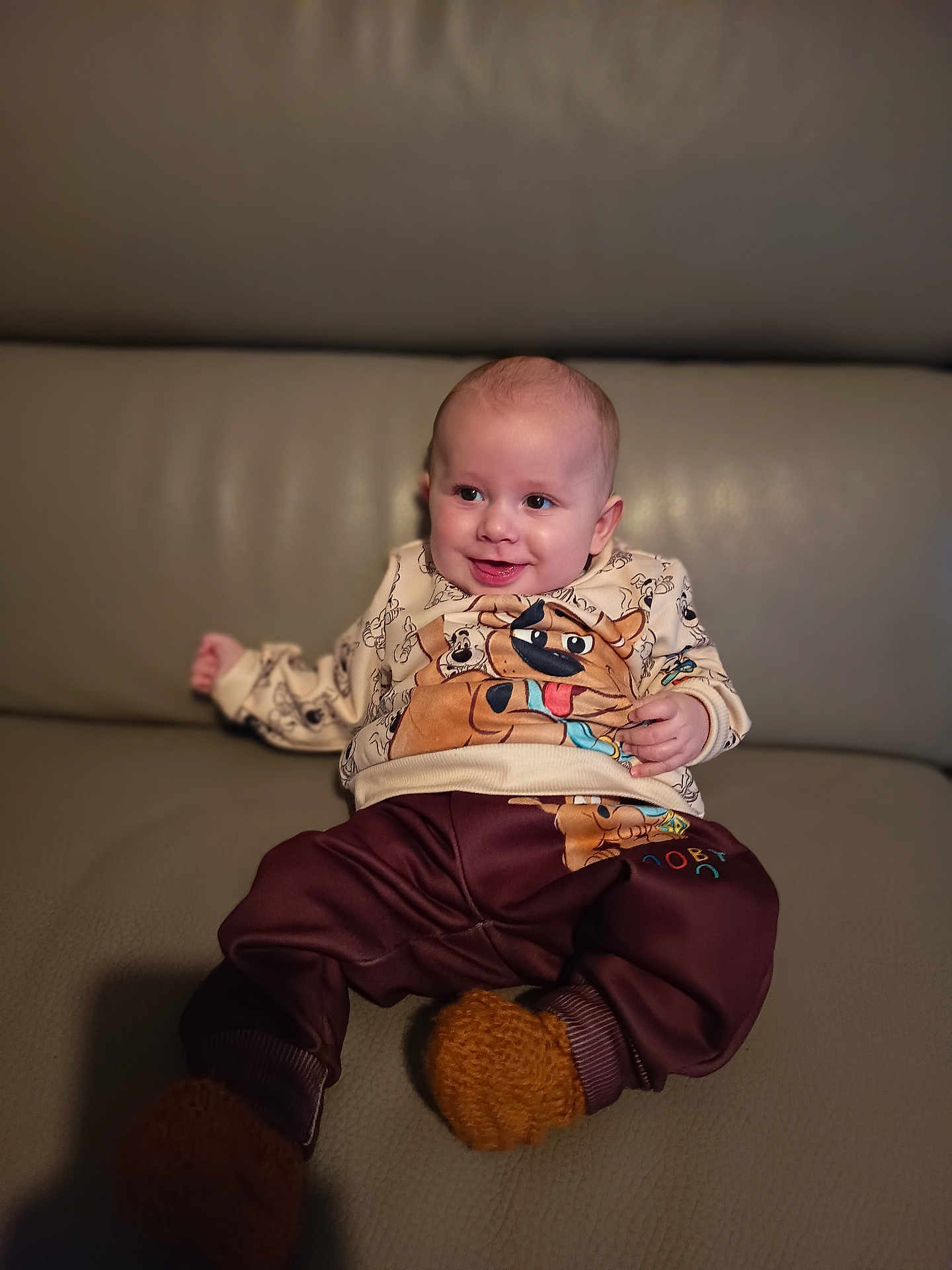 Enoah a rejoint le concours — aidez-le/la à gagner de superbes lots ! baby, infant, smile, sweater, cartoon_print, fuzzy_booties, pants, leather_sofa, couch, portrait, indoor, happy, cute, hands, eyes, head, sitting, skin, bokeh, cozy