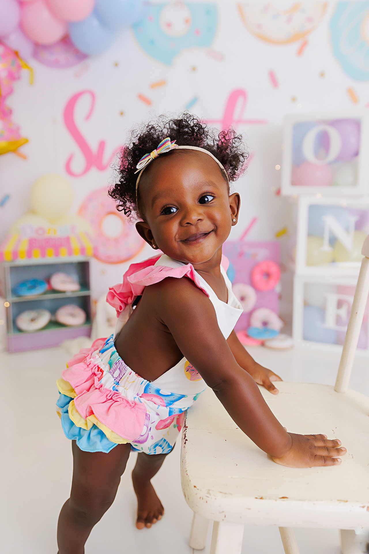 Willow is registered to the contest to win money with this photo: toddler, child, smiling, headband, curly_hair, colorful_clothing, chair, donut_theme, party, balloons, pastel_colors, indoor, happy, cute, celebration, decorations, baby, playful, portrait, person