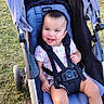 baby, child, stroller, smiling, outdoor, grass, portrait, sitting, harness, safety_belt, infant, happy, face, short_hair, pink_shirt, blue_stroller, park, daylight, wheel, baby_clothing