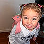 backpack, bright_eyes, casual_clothing, child, collared_shirt, cute, fashion, girl, happy, home, indoor, long_sleeve, person, pink_bows, playful, portrait, smiling, standing, wooden_floor, young_child