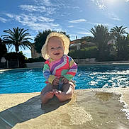 Lena is registered to the contest to win money with this photo: baby, child, pool, water, sunhat, swimsuit, smile, sunshine, outdoor, palm_tree, blue_sky, shadow, concrete, summer, vacation, happy, sitting, reflection, sunlight, nature