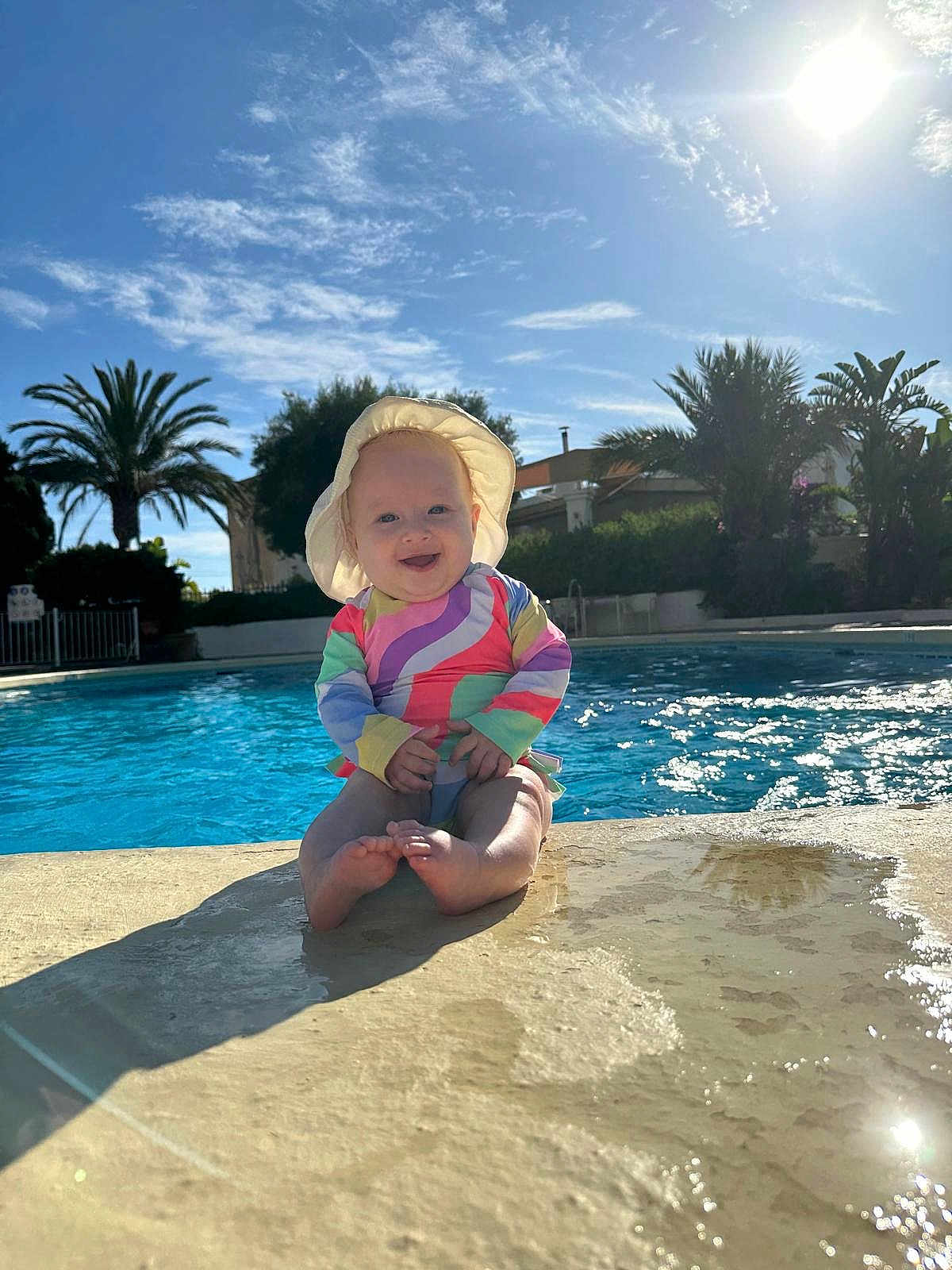 Lena is registered to the contest to win money with this photo: baby, child, pool, water, sunhat, swimsuit, smile, sunshine, outdoor, palm_tree, blue_sky, shadow, concrete, summer, vacation, happy, sitting, reflection, sunlight, nature