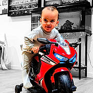 Elyas participe au concours pour gagner de l'argent avec cette photo : toddler, child, toy_motorcycle, red, indoor, floor, training_wheels, smile, face, person, furniture, fish_tank, miniature_motorcycles, shelf, wooden_floor, clothing, long_sleeve, pants, play, adventure