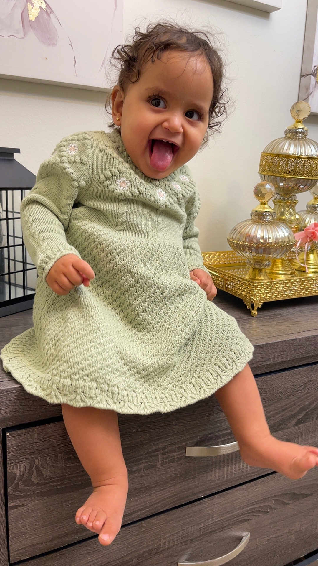 Marya is registered to the contest to win money with this photo: baby, child, curly_hair, cute, decor, earrings, furniture, golden_objects, happy, home, indoor, knitted_dress, person, playful, portrait, sitting, smiling, toddler, tongue_out, wooden_dresser