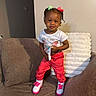 child, toddler, girl, portrait, smiling, standing, indoor, couch, furniture, bows, braids, hair_accessories, t_shirt, red_pants, sneakers, colorful_clothing, living_room, cushions, fashion, photography