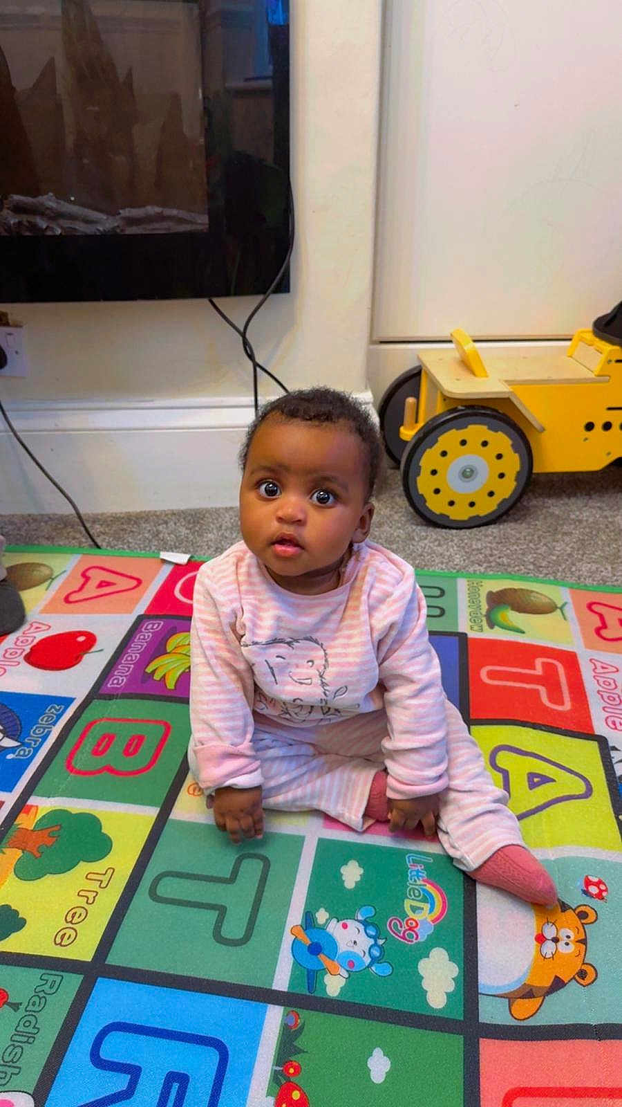 Miracle joined the competition — help win amazing prizes! baby, child, pajamas, play_mat, toy_vehicle, floor, indoor, colorful, alphabet, curious, sitting, carpet, person, infant, cute, young_child, pink_clothing, room, playtime, expression