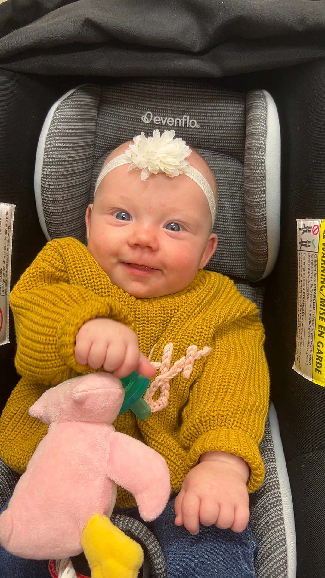 Braelynn is registered to the contest to win money with this photo: baby, child, car_seat, knitted_sweater, yellow_sweater, headband, flower_headband, plush_toy, pink_toy, pacifier, smiling, happy, cute, infant, clothing, indoor, person, seated, blue_jeans, closeup