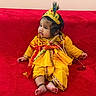 baby, child, infant, traditional_clothing, yellow_outfit, red_background, peacock_feather, crown, headpiece, pearl_garland, cultural_costume, sitting, portrait, indoors, cushion, barefoot, serious_expression, face_paint, colorful, photography