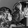affection, animal, black_and_white, child, close_up, couch, curly_hair, dog, face, indoor, interaction, large_dog, nose_to_nose, patterned_cushion, pet, portrait, soft_lighting, tender_moment, wrinkled_face, young_child
