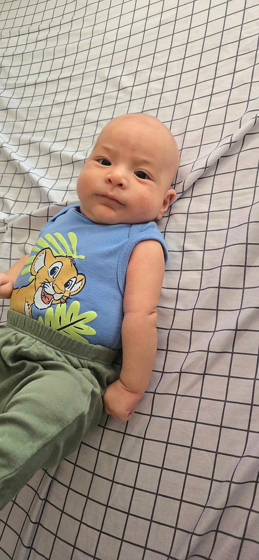 Noah is registered to the contest to win money with this photo: arm, baby, bed, cartoon_print, checkered_pattern, child, clothing, cute, expression, face, indoors, infant, lying_down, pants, person, portrait, sheet, skin, sleeveless_shirt, young