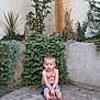 bare_legs, barefoot, brick_wall, casual_clothing, child, concrete_floor, curious_expression, daylight, green_plants, nature, outdoor, plant_leaves, relaxed_pose, sitting, small_child, summer_clothes, sunlight, toddler, urban_garden, young_child
