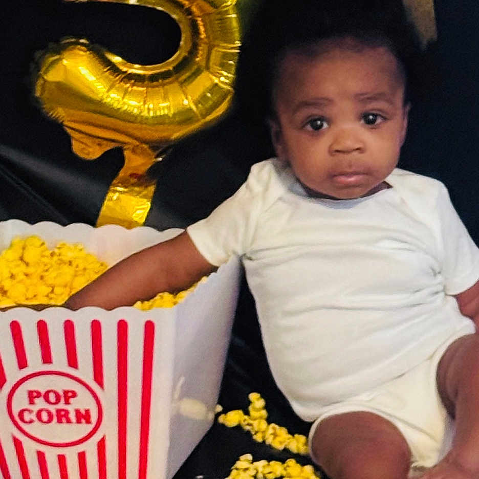 Alijah joined the competition — help win amazing prizes! baby, child, popcorn, balloon, number_5, white_onesie, black_background, sitting, messy, celebration, party, indoor, cute, infant, food, snack, yellow_popcorn, expressionless, floor, event