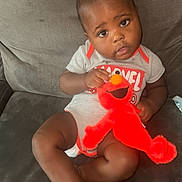 Damani is registered to the contest to win money with this photo: baby, child, plush_toy, elmo, couch, onesie, sitting, curious, indoor, furniture, person, infant, cute, happy, face, toy, clothing, hair, expression, small_hands