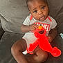 baby, child, plush_toy, elmo, couch, onesie, sitting, curious, indoor, furniture, person, infant, cute, happy, face, toy, clothing, hair, expression, small_hands