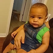 Malachi is registered to the contest to win money with this photo: toddler, child, indoor, denim_overalls, green_shirt, person, sitting, hand, face, expressionless, furniture, door, hallway, floor, tile_floor, adult, couch, skin, casual_clothing, home
