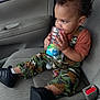 toddler, child, car_interior, car_seat, curly_hair, camo_pants, black_shoes, water_bottle, chewing, seatbelt, car_door, window, plastic_bottle, young_child, sitting, casual_clothing, side_view, indoors, baby, person
