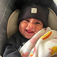 Eliana joined the competition — help win amazing prizes! baby, smiling, blanket, hat, black_clothing, car_seat, face, child, cute, cozy, indoor, infant, headwear, happy, person, portrait, warm, childcare, closeup, comfort