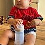 Kylian participe au concours pour gagner de l'argent avec cette photo : toddler, child, baby_bottle, bread, red_shirt, hugo_boss, blue_sneakers, socks, wooden_table, indoor, curly_hair, cute, sitting, food, drink, casual_clothing, young_child, face, legs, hand