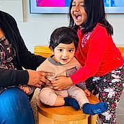 Ishaan Kuna is registered to the contest to win money with this photo: child, baby, girl, laughing, smiling, indoor, stool, clothing, red_shirt, patterned_leggings, adult_hand, supporting, curly_hair, short_hair, wooden_furniture, carpet, screen, colorful_background, happy, family