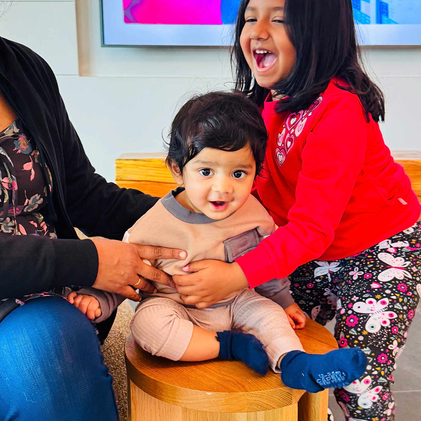Ishaan Kuna is registered to the contest to win money with this photo: child, baby, girl, laughing, smiling, indoor, stool, clothing, red_shirt, patterned_leggings, adult_hand, supporting, curly_hair, short_hair, wooden_furniture, carpet, screen, colorful_background, happy, family