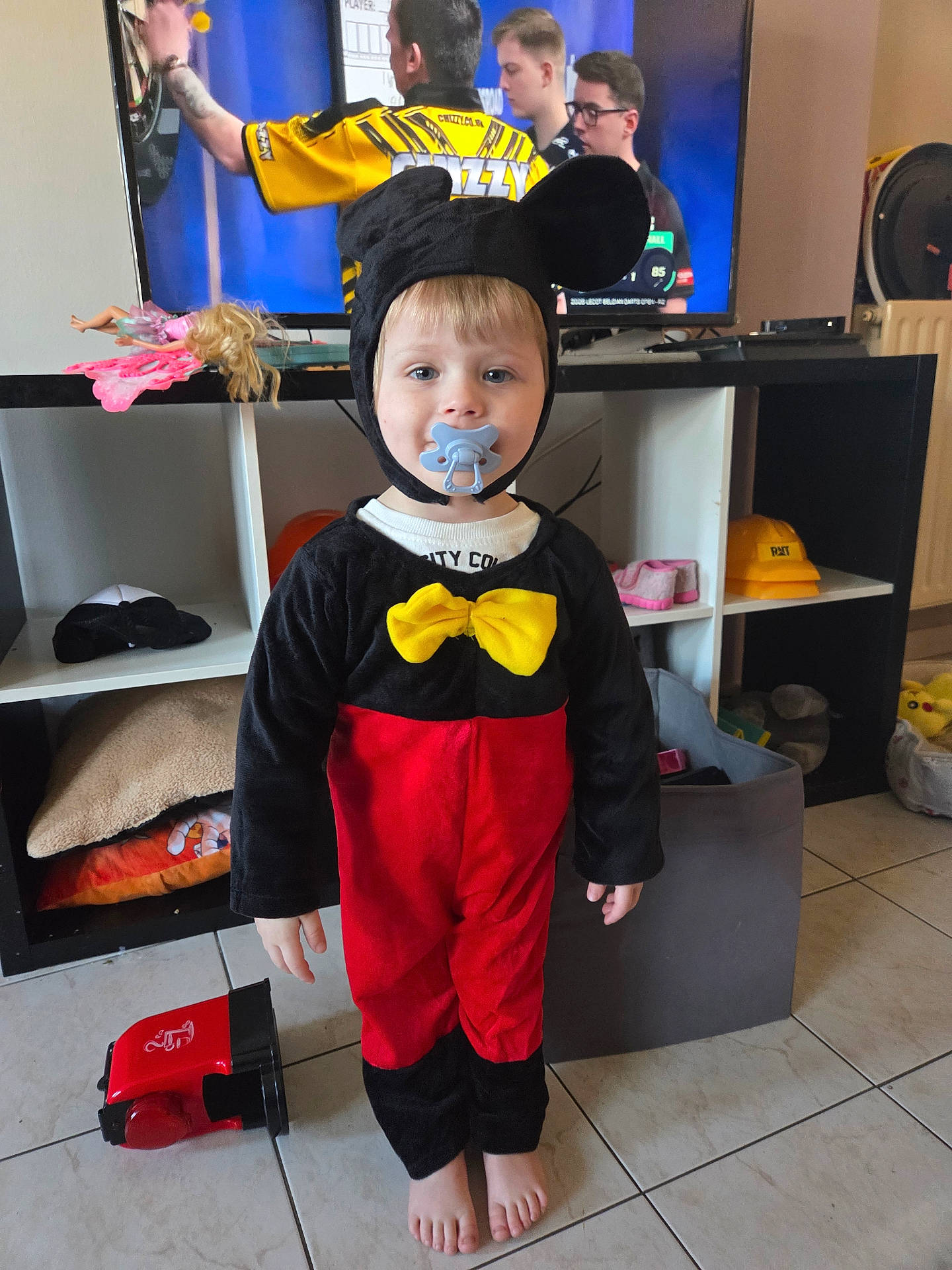 Sohan a rejoint le concours — aidez-le/la à gagner de superbes lots ! barefoot, black_red_outfit, child, costume, darts, floor, furniture, glasses, hat, home, indoor, mickey_mouse, pacifier, person, shelf, television, tile, toddler, toy, yellow_bow