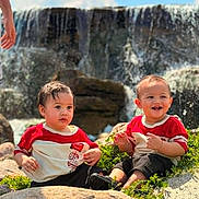 Jeraiah joined the competition — help win amazing prizes! toddler, child, waterfall, rocks, greenery, outdoor, nature, smiling, happy, sitting, red_shirt, shorts, sunny, daylight, grass, cute, two_children, water, playful, portrait