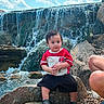 Jeraiah joined the competition — help win amazing prizes! toddler, child, waterfall, rock, outdoor, nature, sky, clouds, water, person, red_shirt, black_shorts, footwear, rocks, pebbles, casual, summer, daytime, curious, family