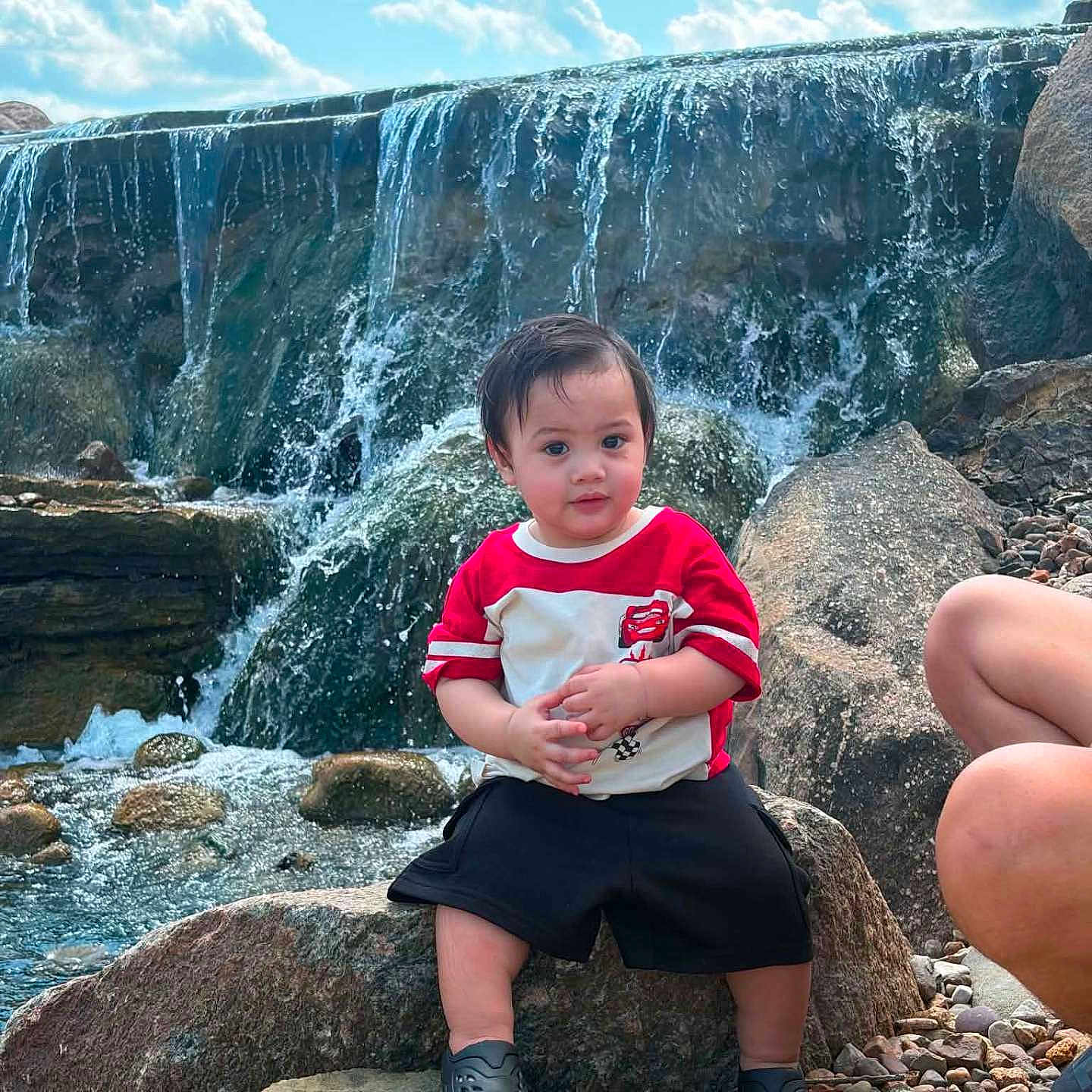Jeraiah joined the competition — help win amazing prizes! toddler, child, waterfall, rock, outdoor, nature, sky, clouds, water, person, red_shirt, black_shorts, footwear, rocks, pebbles, casual, summer, daytime, curious, family
