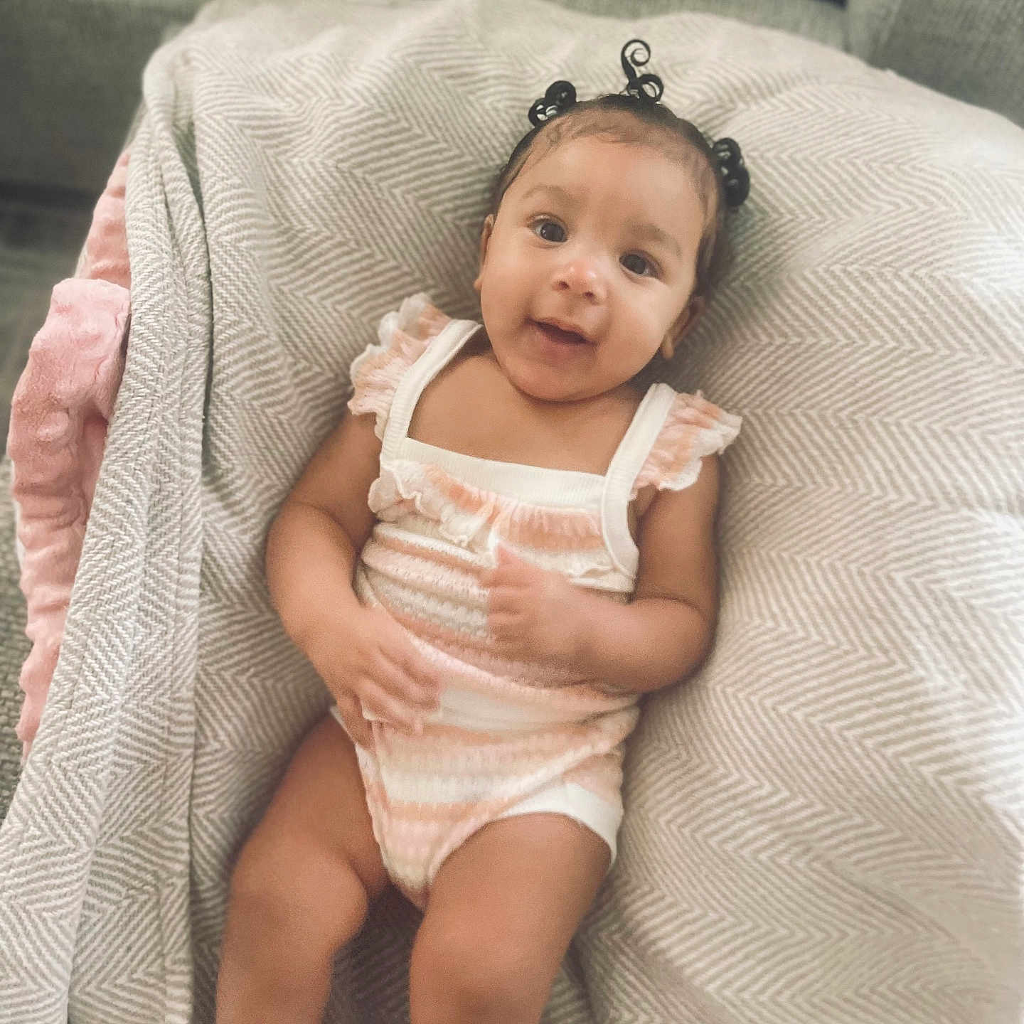 Mauvi is registered to the contest to win money with this photo: baby, blanket, child, clothing, comfort, cozy, cute, face, hair_buns, happy, indoors, infant, onesie, patterned_blanket, person, portrait, sitting, skin, smile, soft_fabric