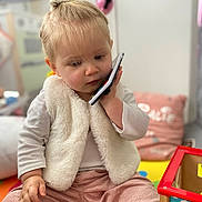 Calie participe au concours pour gagner de l'argent avec cette photo : toddler, child, phone, indoor, toy, pink_pants, white_vest, hair_bow, play_area, cushion, floor_mat, curious, sitting, young_child, person, soft_lighting, corduroy, holding_phone, colorful, casual_clothing