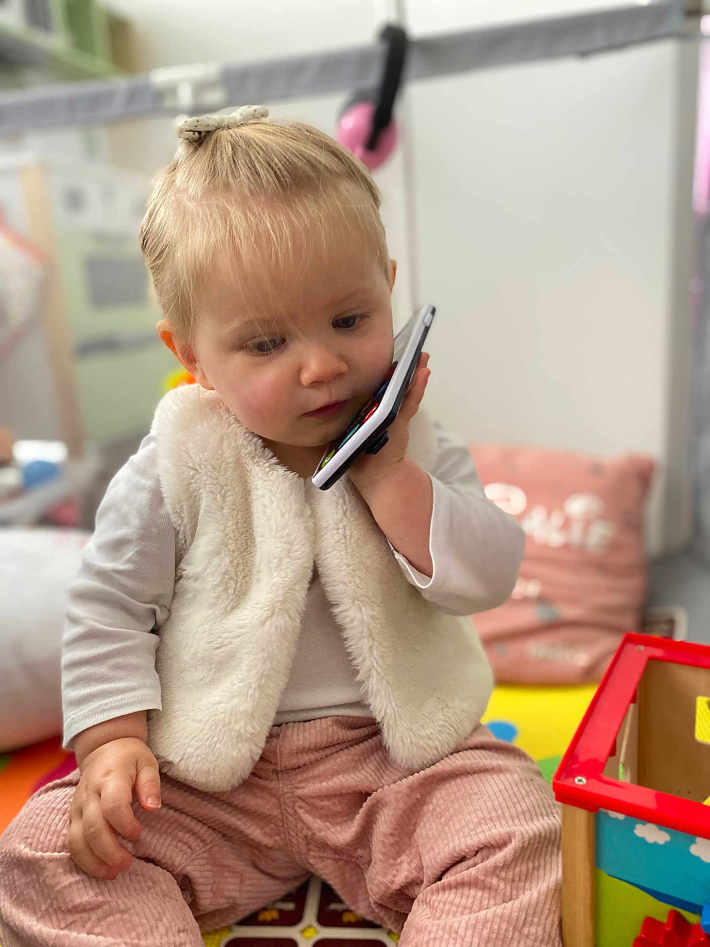 Calie participe au concours pour gagner de l'argent avec cette photo : toddler, child, phone, indoor, toy, pink_pants, white_vest, hair_bow, play_area, cushion, floor_mat, curious, sitting, young_child, person, soft_lighting, corduroy, holding_phone, colorful, casual_clothing