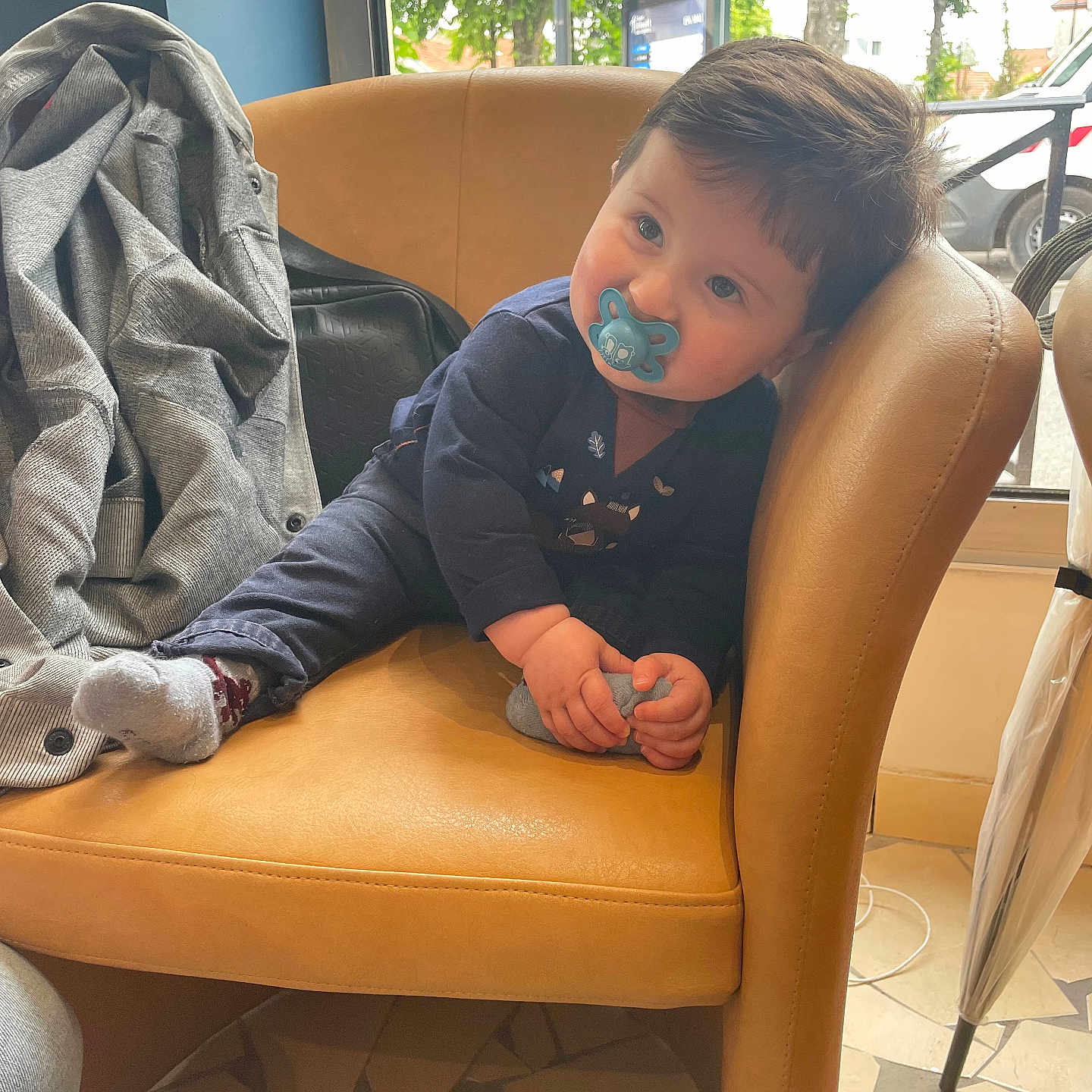 Joris participe au concours pour gagner de l'argent avec cette photo : baby, candid, casual_clothing, chair, child, curious, daylight, floor, foot, holding, home_interior, indoor, jacket, leather, pacifier, person, relaxed, seated, window, young_child