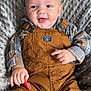 baby, child, smiling, blue_eyes, overalls, plaid_shirt, toy, cushion, indoor, seated, happy, face, hands, clothing, texture, cute, infant, person, portrait, comfortable