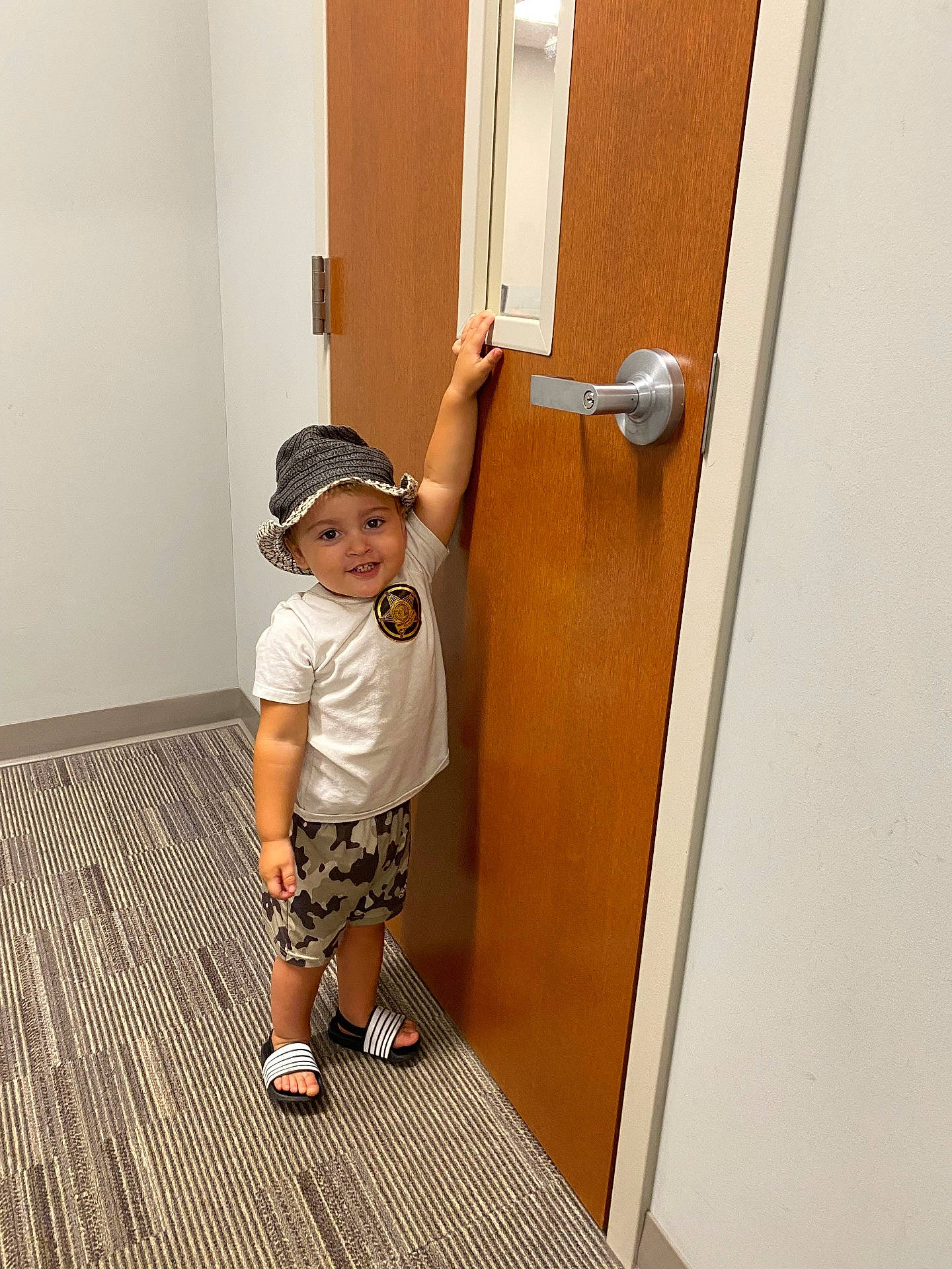 boy, camouflage, carpet, child, door, expression, footwear, hand, handle, hat, indoor, person, sandals, shirt, shorts, smile, standing, toddler, wall, young_child