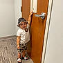 badge, carpet, child, door, door_handle, floor, footwear, hand, hat, indoor, pattern, person, shorts, smile, standing, striped, toddler, tshirt, wall, wood