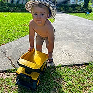 Lathan joined the competition — help win amazing prizes! toddler, child, toy, dump_truck, yellow, hat, shorts, bare_chest, grass, sidewalk, driveway, house, brick_house, front_porch, greenery, sunlight, outdoor, playing, curious, summer