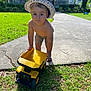 toddler, child, toy, dump_truck, yellow, hat, shorts, bare_chest, grass, sidewalk, driveway, house, brick_house, front_porch, greenery, sunlight, outdoor, playing, curious, summer