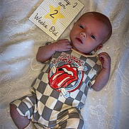Creed is registered to the contest to win money with this photo: baby, infant, blanket, onesie, rolling_stones_logo, milestone_card, text, lying_down, socks, cute, newborn, soft_texture, white, patterned_clothing, young_child, portrait, face, hand, foot, sleepwear