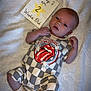 baby, infant, blanket, onesie, rolling_stones_logo, milestone_card, text, lying_down, socks, cute, newborn, soft_texture, white, patterned_clothing, young_child, portrait, face, hand, foot, sleepwear