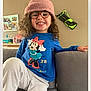 child, smiling, glasses, hat, curly_hair, blue_shirt, minnie_mouse, pink_hat, couch, living_room, cozy, happy, person, indoor, casual_clothing, pajama_pants, home, furniture, portrait, cute