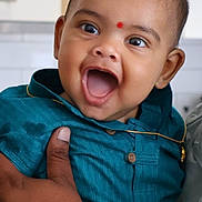 Arush joined the competition — help win amazing prizes! baby, child, smiling, happy, face, person, hand, clothing, blue_shirt, indoor, portrait, cute, infant, gold_chain, red_mark, skin, eyes, mouth, holding, adult_hand