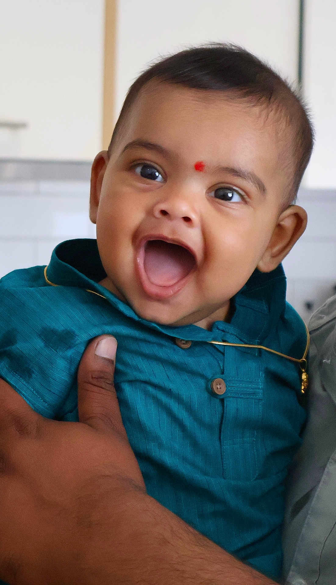 Arush joined the competition — help win amazing prizes! baby, child, smiling, happy, face, person, hand, clothing, blue_shirt, indoor, portrait, cute, infant, gold_chain, red_mark, skin, eyes, mouth, holding, adult_hand